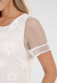 Femme portant un haut en dentelle blanche avec broderie florale et manches courtes en mesh transparent ornées d'un galon décoratif.