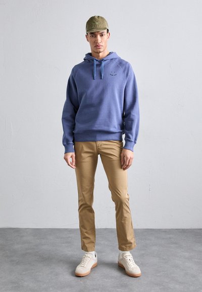 Sweatshirt com capuz em azul claro com cordão em contraste, chinos bege e ténis brancos com solas e atacadores em bege. Boné verde-oliva na cabeça.