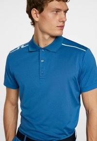 Blå polo skjorta i glatt syntetmaterial, med krage, tre-knapp placket och vita detaljer på axlarna.