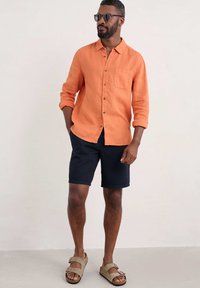 Oranje linnen overhemd met knoopsluiting en een borstzak, gecombineerd met marineblauwe shorts en beige sandalen. Zonnebril voegt een moderne accent toe.