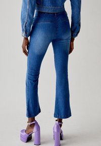 Jeans flare in denim blu con vita media, dotati di tasche posteriori e una texture liscia. Abbinati a tacchi a piattaforma viola.