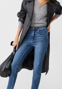 Musta nahkainen trenssitakki vaaleanharmaan svetarin päällä, yhdistetty sinisiin korkeavyötäröisiin farkkuihin. Asussa on jäsennelty siluetti ja sileät tekstuurit.