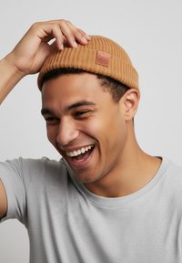 Tan knitted beanie with ribbed texture and a leather tag. Gray t-shirt visible. Subject shows a joyful expression while adjusting the hat.