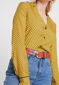 Gul skjorte med knapper og sorte trekantede mønstre, lange ærmer og krave. Sammen med højtaljede blå denimbukser og et rødt tekstureret bælte.