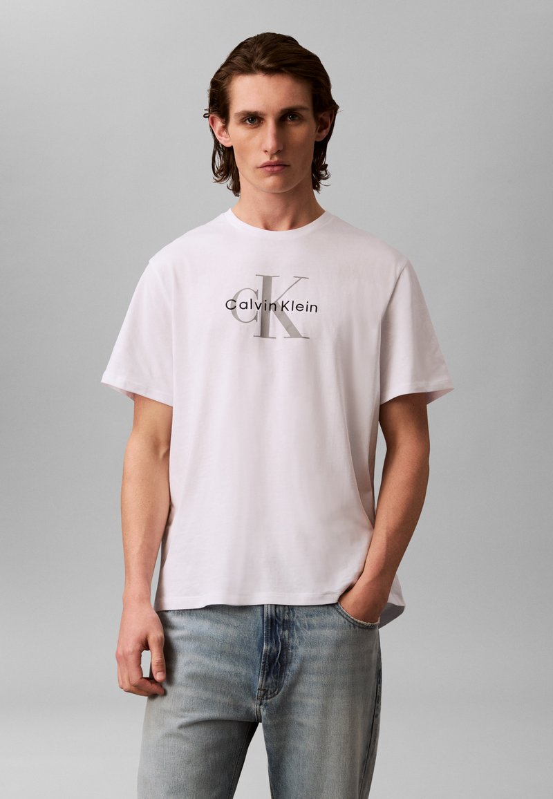 Fiatal férfi középhosszú barna hajjal, fehér Calvin Klein pólót és világoskék farmert visel, áll, egyik kezét zsebre téve.