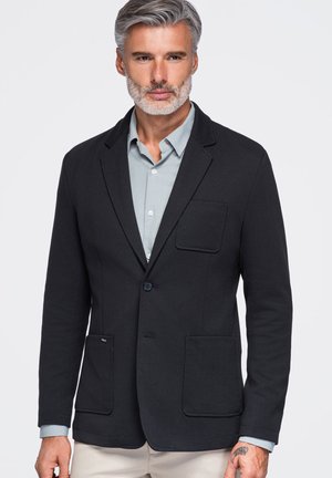 Hombre con cabello y barba gris que lleva una chaqueta negra sobre una camisa azul clara abotonada con fondo liso.