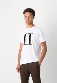 Camiseta de algodón blanca con una gran impresión negra de "II" en la parte frontal. Mangas cortas y escote redondo. Combinada con pantalones marrones.