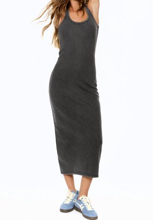 Femme portant une robe mi-longue sans manches gris foncé associée à des baskets bleu clair et blanc, debout devant un fond blanc.