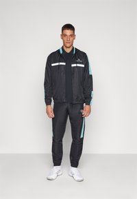 Hombre vestido con un chándal deportivo negro y verde azulado, con zapatillas blancas, de pie frente a un fondo gris claro liso.