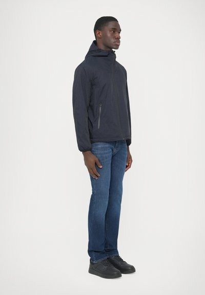 Námornícky vodeodolná bunda s vreckom na zips na boku, doplnená modrými džínsami a čiernymi teniskami. Má kapucňu a elastické manžety.