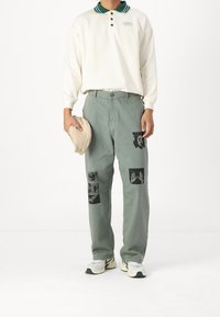 Osoba w białej koszuli polo z długim rękawem i zielonym kołnierzykiem, szarych spodniach z czarnymi nadrukami graficznymi, białych trampkach, trzymająca beżowy kapelusz w lewej ręce.