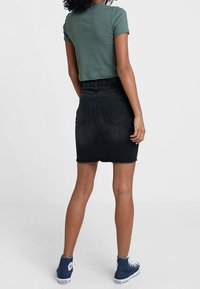 Svart denimkjol med råkant, kombinerad med en grön ribbad topp med kort ärm och blå hög-top sneakers, sedd bakifrån.