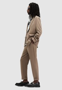 Traje marrón a medida con un ajuste estructurado, combinado con una camisa de diseño y zapatos negros robustos. El modelo tiene largas rastas negras y gafas de sol.