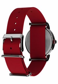 Correa de reloj de tela roja con herrajes de metal en tono plateado, que presenta una textura suave y una hebilla ajustable. Caja de reloj redonda visible en el lateral.