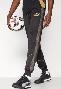 Musta jogger-housut kultaisilla yksityiskohdilla ja kuvioidulla sivuraidalla. Käytetty mustien jalkapallokenkien kanssa ja pitelee mustavalkoista jalkapalloa.