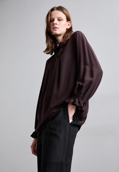 Blusa marrone, fluida, con maniche lunghe e polsi arricciati; presenta un colletto a oblò e un tessuto morbido; indossata sopra pantaloni neri.