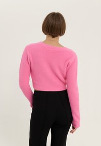Cropad tröja i mjuk rosa tyg med texturerad stickning, långa ärmar och vid halsringning, tillsammans med svarta åtsittande byxor.