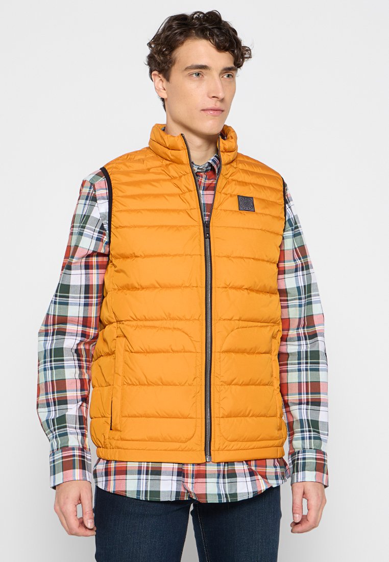 Gilet rembourré orange avec un col montant et une fermeture éclair, porté sur une chemise en flanelle à carreaux multicolores, dotée de poches latérales et d'un patch logo.