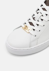 Sneaker bianco in pelle con punta rotonda, dettagli marroni testurizzati, lacci bianchi piatti e un logo dorato sul porta-lacci.