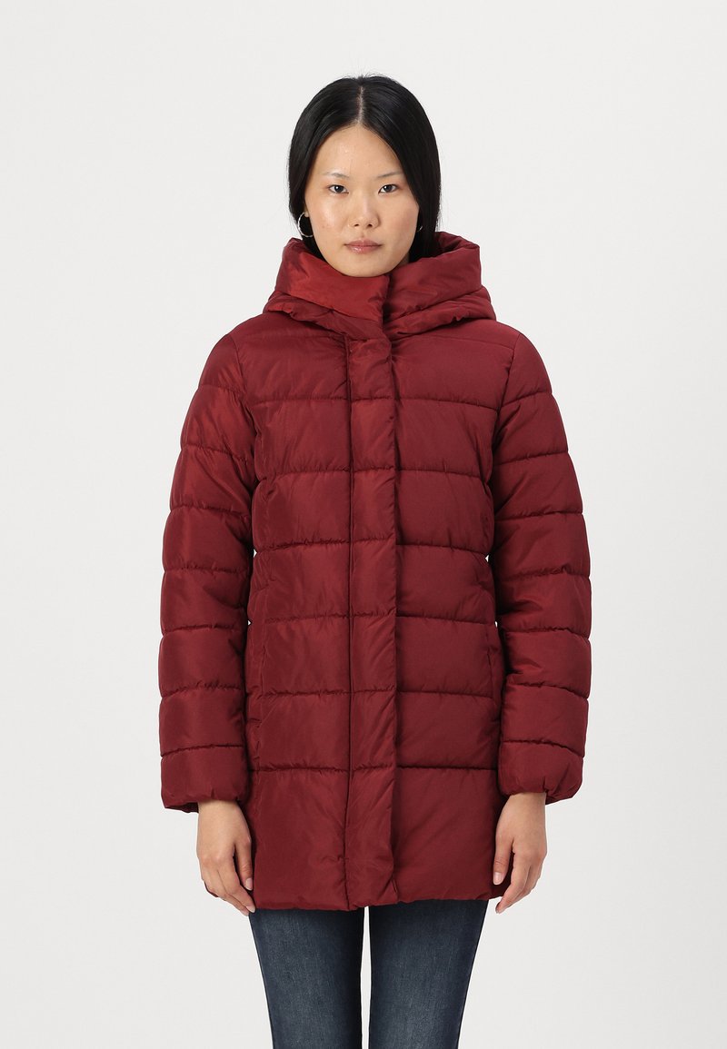 Manteau matelassé bordeaux avec un col haut et une capuche, présentant un design matelassé et une fermeture éclair sur toute la longueur, associé à un jean en denim foncé.
