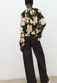 Blusa nera fluida con grande stampa floreale crema, scollatura arricciata e maniche lunghe, abbinata a pantaloni a gamba larga marroni scuri a vita alta.