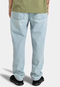 Pantalon en denim bleu clair avec une coupe droite, comprenant deux poches arrière et une texture subtilement délavée. Porté avec des chaussures noires.