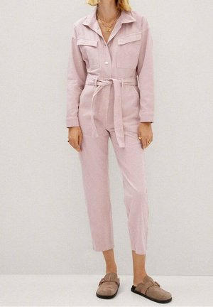 Nainen pukeutuneena vaaleanpunaiseen vyölliseen haalariasuun, jossa on pitkät hihat ja lyhyt lahje, yhdistettynä beigen värisiin liukusolkuihin.