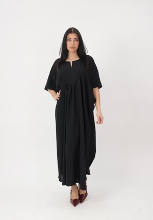 Femme aux longs cheveux noirs portant une robe maxi noire ample et des talons pointus bordeaux, debout devant un fond blanc.