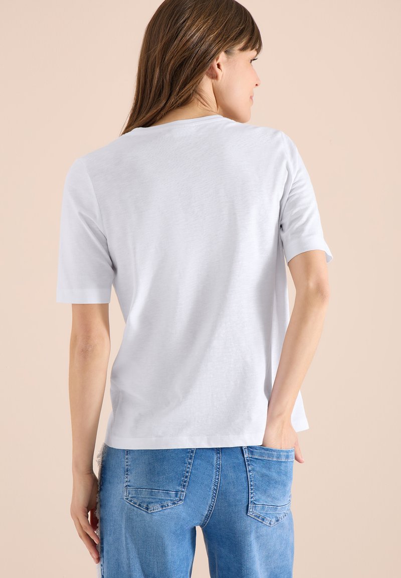 Femme vue de dos portant un t-shirt blanc uni à manches courtes et un jean bleu, avec une main dans la poche arrière.
