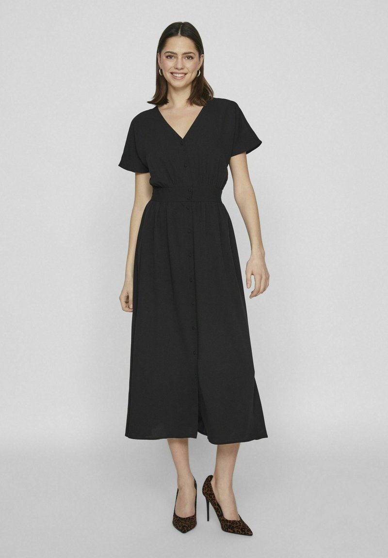 Čierne šaty strednej dĺžky s krátkymi rukávmi, výstrihom do V a zapínaním na gombíky. Ľahký materiál s zúženým pásom a splývavou sukňou. Topánky s leopardím vzorom.