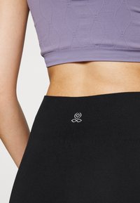 Mustat legginssit, joissa on sileä pinta ja tiukka vyötärö, taustalla pieni valkoinen brodeerattu logo. Yllä violetti tekstuurinen crop-top.