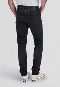 Mann trägt schwarze Slim-Fit-Jeans und weiße Sneaker, von hinten gezeigt mit den Händen auf den Hüften vor schlichtem Hintergrund.