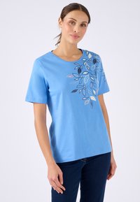 Mujer vestida con una camiseta de manga corta azul claro con un diseño de hojas multicolores en el hombro, combinada con jeans azul oscuro, de pie frente a un fondo liso.