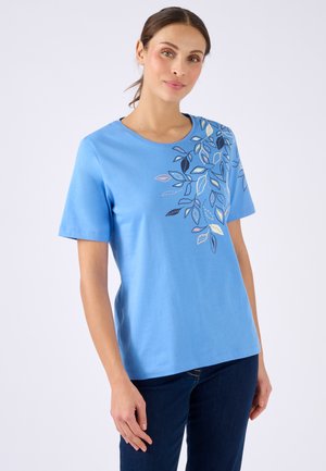 Vrouw in een lichtblauw shirt met korte mouwen en een meerkleurig bladerenontwerp op de schouder, gecombineerd met donkerblauwe spijkerbroek, staande tegen een effen achtergrond.