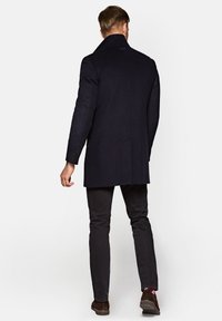 Granatowy płaszcz z mieszanki wełny z wyraźnym kołnierzem, pojedynczym wentylem z tyłu i dopasowanym krojem. Noszony na ciemnych spodniach i brązowych butach.