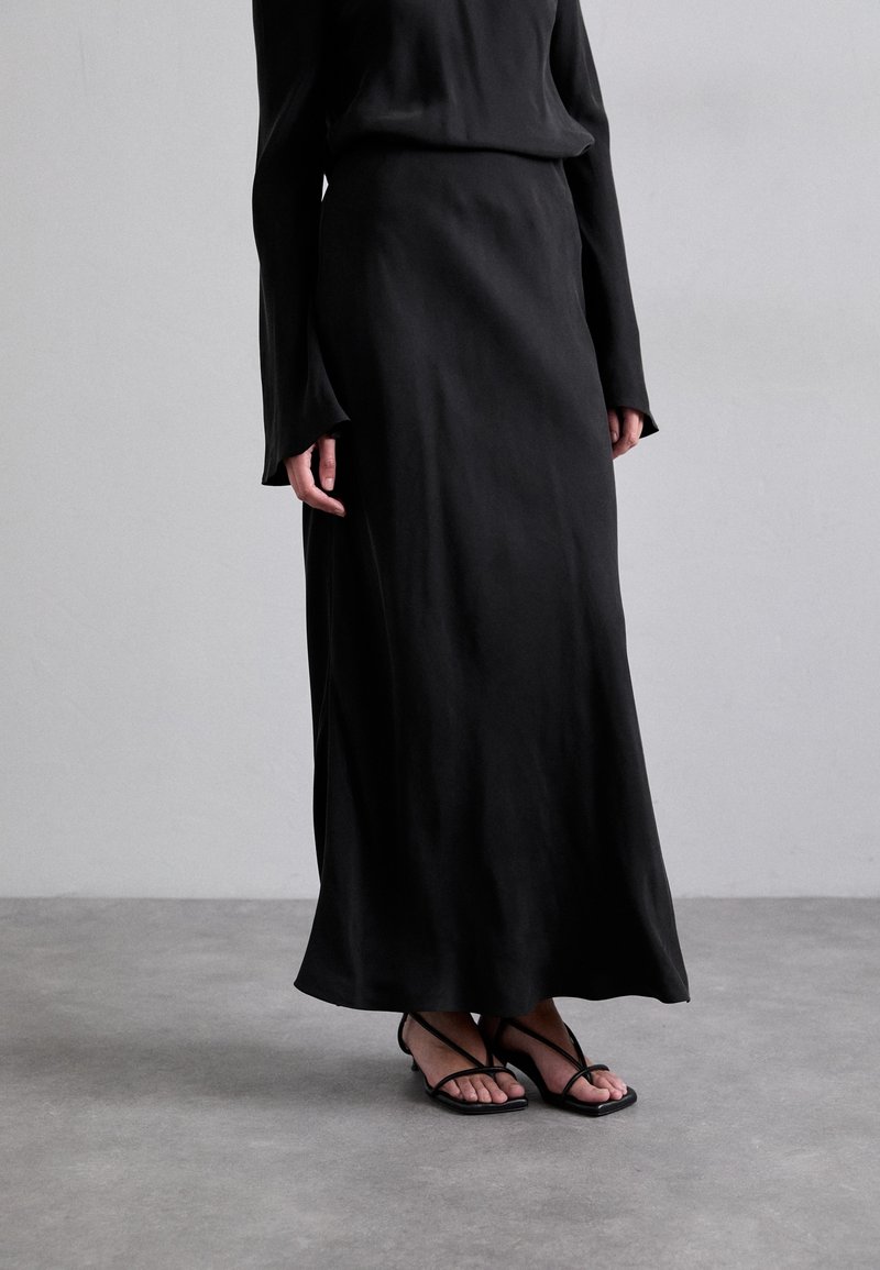 Personne portant une longue jupe noire, un haut noir à manches longues et des sandales noires à brides et orteils ouverts, debout sur un sol gris.