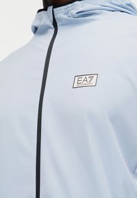 Ανοιχτό μπλε μπουφάν με μαύρο φερμουάρ και το λογότυπο EA7 Emporio Armani στο στήθος, φορεμένο με την κουκούλα σηκωμένη.