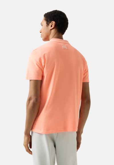 Muž otočený zády, má na sobě lososově oranžové polo tričko s krátkými rukávy a světle šedé kalhoty, na pozadí je čistě bílá stěna.