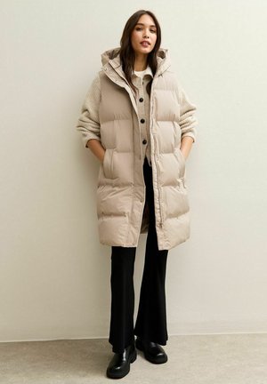Chaleco acolchado beige con capucha, con botones a presión y bolsillos laterales, llevado sobre un suéter ligero y pantalones acampanados negros, complementado con botas negras.