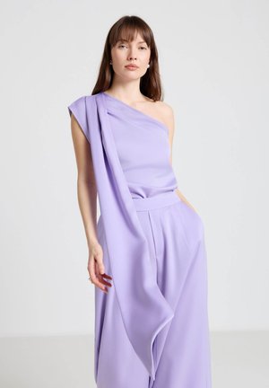 Femme portant une combinaison lavande fluide à une épaule avec des jambes larges, debout devant un fond uni gris clair.