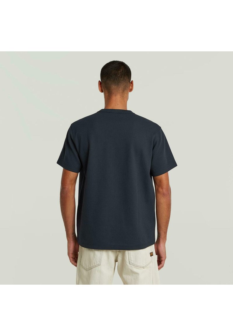 Hombre con camiseta oscura de manga corta y pantalones claros, de pie de espaldas sobre un fondo neutro.