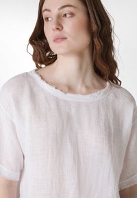 Camicia bianca di lino con scollatura ampia, maniche corte e bordi sfilacciati. Il tessuto ha un aspetto leggero e strutturato.