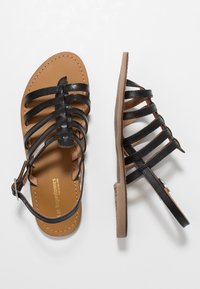 Sandalias negras de tiras con suelas planas, que cuentan con múltiples tiras de cuero y una hebilla ajustable en el tobillo. Plantilla de color marrón claro con detalle de costura.