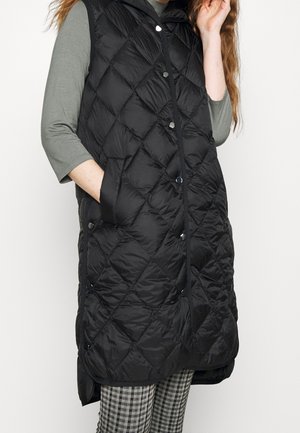 Femme portant un gilet matelassé noir sans manches avec des boutons-pression par-dessus une chemise verte à manches longues et un pantalon à carreaux noir et blanc.