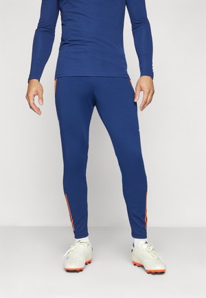 Homme portant des leggings de sport bleus ajustés et un haut à manches longues, avec des crampons blancs à semelles orange, debout sur un fond uni.