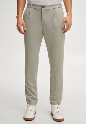 Mężczyzna ubrany w jasnoszare teksturowane spodnie i białe trampki, stojący z rozluźnionymi rękami wzdłuż ciała na białym tle.