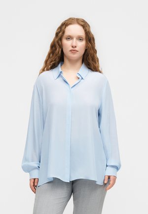 Frau mit langen lockigen Haaren, die eine locker sitzende hellblaue Bluse mit Knöpfen und hellgraue Hosen trägt, steht vor einem einfarbigen weißen Hintergrund.