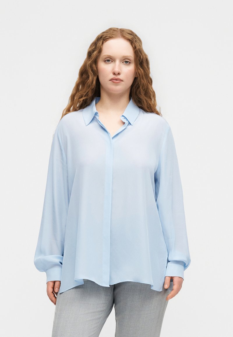 Frau mit langen lockigen Haaren, die eine locker sitzende hellblaue Bluse mit Knöpfen und hellgraue Hosen trägt, steht vor einem einfarbigen weißen Hintergrund.