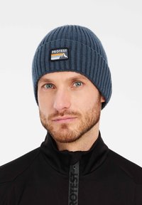 Cappello in maglia a coste blu con un imponente patch nero e multicolore "PROTEST" che presenta grafiche di montagne. Tessuto morbido, design aderente.