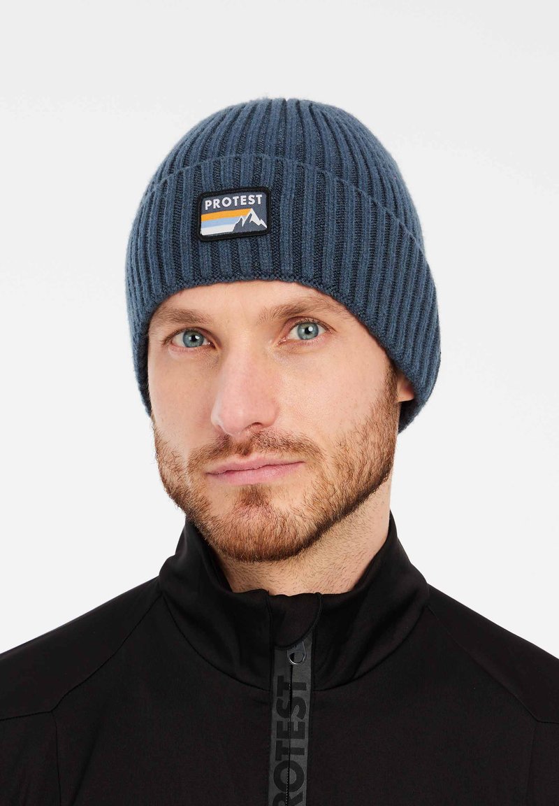 Cappello in maglia a coste blu con un imponente patch nero e multicolore "PROTEST" che presenta grafiche di montagne. Tessuto morbido, design aderente.